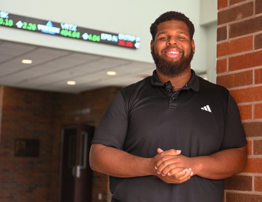 A male student standing in the College of Business lobby in front of the stock ticker