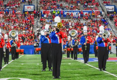 Marching band members in blue and red uniforms perform with brass instruments on a football field, with a packed stadium of fans in the background