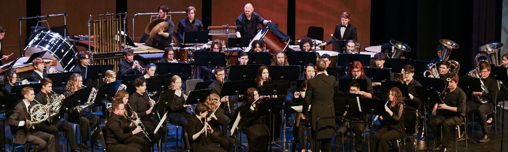 A large university concert band performs on stage, with musicians playing woodwinds, brass, and percussion under the direction of a conductor.