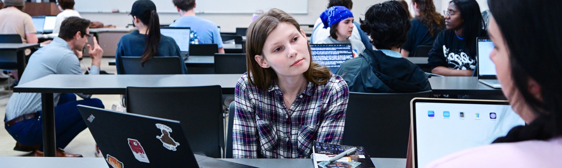 Students sit at tables in a classroom working on laptops, with a student in a plaid shirt listening attentively during a small group discussion in the foreground.