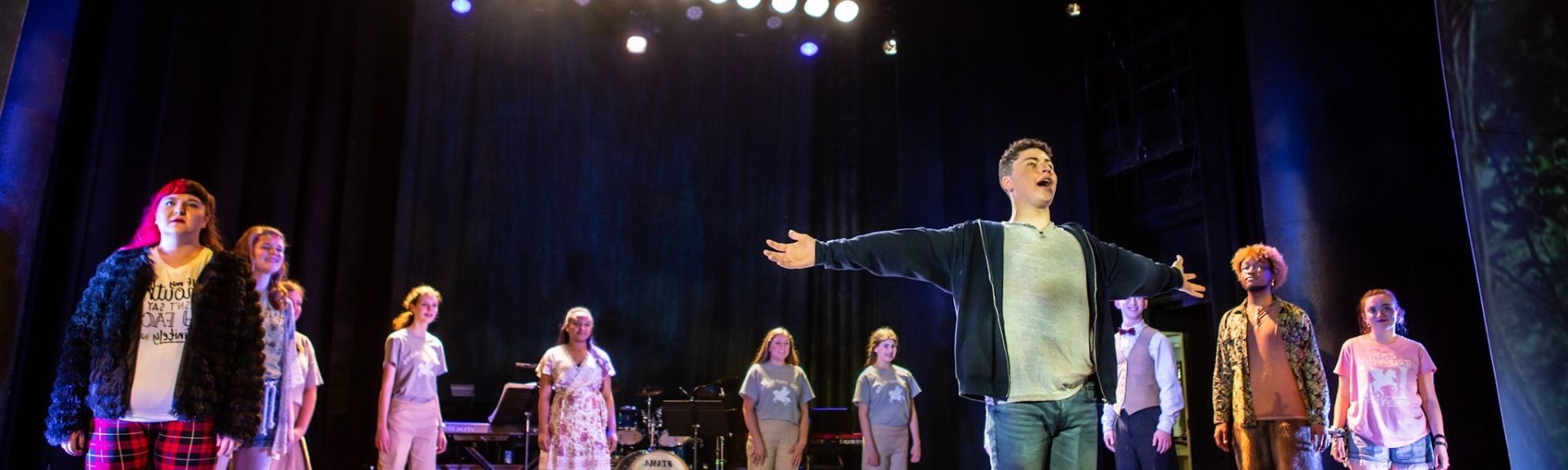 A group performs on a dimly lit theater stage, with one person center stage arms wide open, lit by stage lights and a dark backdrop. Musical instruments sit in the background.
