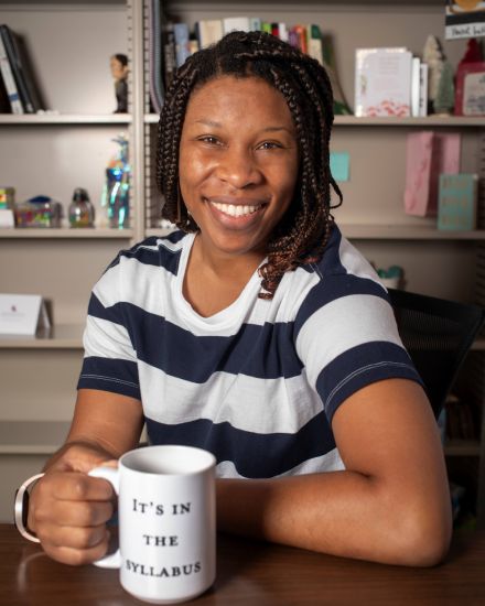 Alana Wagner sitting in her office holding a mug that says "it's in the syllabus"